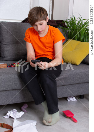Child sitting on sofa in casual clothes, focused on smartphone while surrounded by toys and clothes Child sitting on sofa in casual clothes, focused on smartphone while surrounded by toys and clothes 138833594