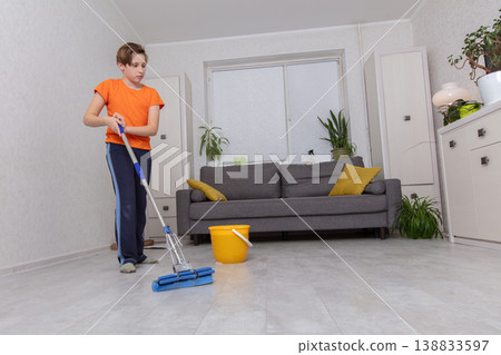 Young boy cleans living room floor with mop and bucket in bright, modern home during daytime Young boy cleans living room floor with mop and bucket in bright, modern home during daytime 138833597