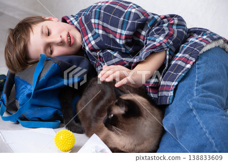 Young boy relaxes with a cat while lying on the floor beside his backpack and colorful toys at home in the afternoon Young boy relaxes with a cat while lying on the floor beside his backpack and colorful toys at home in the afternoon 138833609