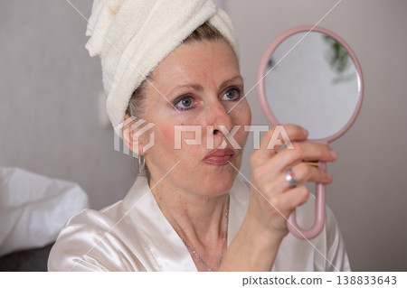 Woman applies skincare while looking in a handheld mirror at home in a cozy setting Woman applies skincare while looking in a handheld mirror at home in a cozy setting 138833643