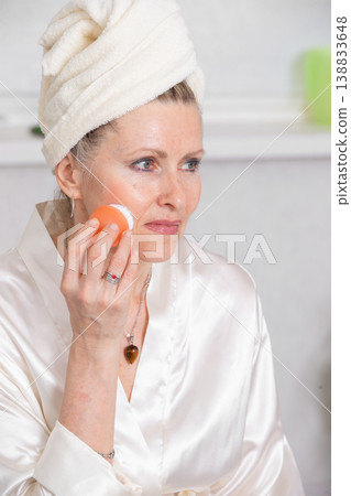 Woman applying skincare treatment in a spa-like setting while wearing a silk robe and towel on her head Woman applying skincare treatment in a spa-like setting while wearing a silk robe and towel on her head 138833648