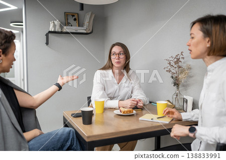 Women sitting at table eating and talking in breakfast kitchen area of open office. Eating, table, kitchen, teamwork, business, women. 138833991