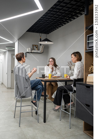 Women eating and talking at office kitchen table with colleagues during lunch break. Eating, talking, kitchen, office, teamwork, women. Women eating and talking at office kitchen table with colleagues during lunch break. Eating, talking, kitchen, office, teamwork, women. 138833992