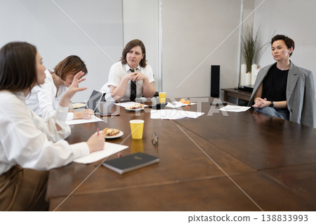 Team of women at table working and eating during morning office meeting with cups and croissants. Morning, team, eating, table, office, croissants, women. 138833993