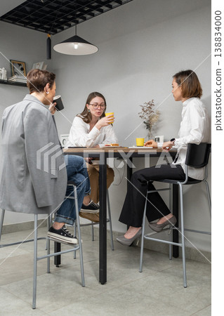 Three women gathered around high table in kitchen area chatting and eating during office lunch. Eating, chatting, kitchen, teamwork, lunch, office, women. 138834000