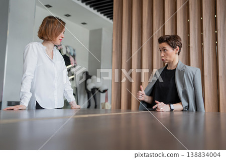 Two women standing in office at desk, talking and exchanging ideas in professional environment. Talking, ideas, office, women, teamwork. 138834004