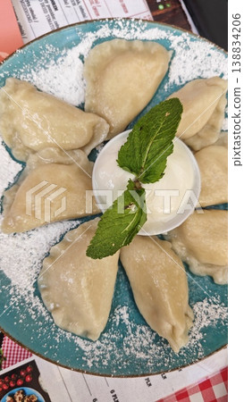 Pierogi served on a turquoise plate, arranged in a circular formation with a bowl of cream and fresh mint in the center, dusted with powdered sugar Pierogi served on a turquoise plate, arranged in a circular formation with a bowl of cream and fresh mint in the center, dusted with powdered sugar 138834206