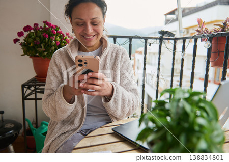 Smiling Woman Using Smartphone on Cozy Balcony Surrounded by Plants and Laptop 138834801