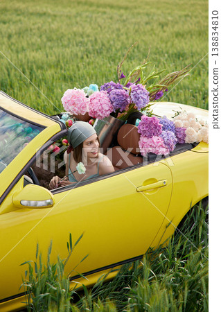 Smiling woman sits in yellow convertible surrounded by colorful flowers Smiling woman sits in yellow convertible surrounded by colorful flowers 138834810