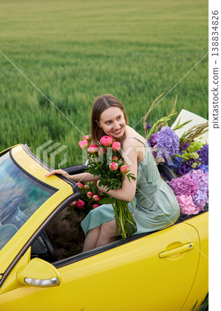 Smiling woman sits in yellow convertible surrounded by colorful flowers 138834826