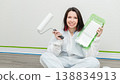 Woman in protective coveralls holds paint roller and tray while sitting on the floor of a freshly painted room with green tape on the walls 138834913