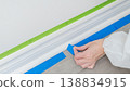 Person applying blue painter's tape along baseboard in preparation for painting, with green tape visible on the wall above 138834915