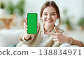 Young woman holds smartphone with green screen, smiling and pointing in a bright indoor setting with plants and furniture in the background 138834971