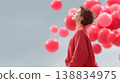 Woman in red sweater stands with eyes closed, surrounded by floating red balloons against a soft gray background 138834975