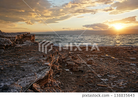Sun shines through dramatic sunset clouds to waves at Jordanian side of Dead Sea, branches, flotsam, debris and rubbish washed on shore 138835643