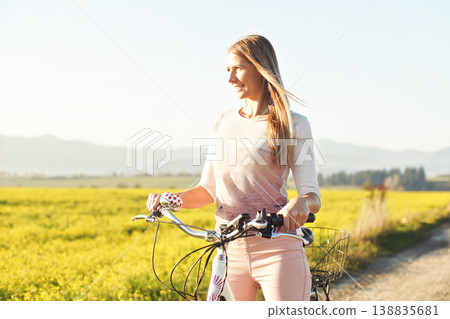 Young woman standing next to her bike over dusty country road, strong afternoon sun backlight in background shines on yellow flowers field 138835681
