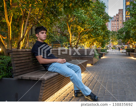 New york, usa august 15,2024. Young man sitting on a high line bench at golden hour, relaxed and thoughtful amid manhattan trees, architecture and evening light 138835692
