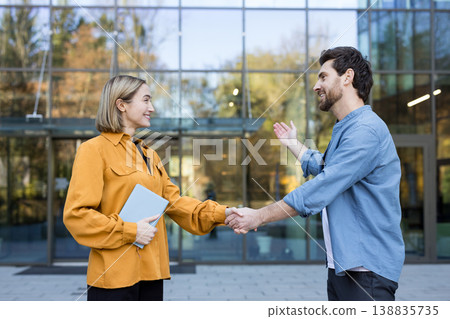 Two smiling business people shaking hands in front of a modern glass building, signifying a successful agreement, teamwork, and a new professional relationship 138835735