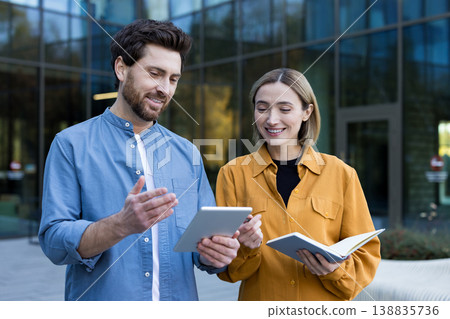 Business professionals engaging in an informal outdoor meeting, discussing work and ideas using a digital tablet and notebook in front of a modern office building Business professionals engaging in an informal outdoor meeting, discussing work and ideas using a digital tablet and notebook in front of a modern office building 138835736