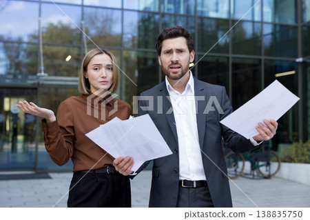 Confused business people standing outdoors in front of an office building, holding documents and shrugging, expressing frustration, misunderstanding, failure, and stress in their work 138835750