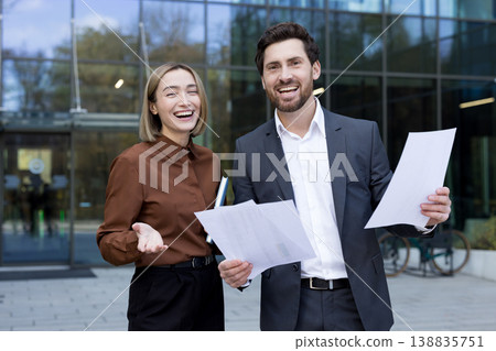 Two cheerful business professionals, man and woman, standing outside a modern corporate building, holding documents and smiling, conveying teamwork, success and confident collaboration Two cheerful business professionals, man and woman, standing outside a modern corporate building, holding documents and smiling, conveying teamwork, success and confident collaboration 138835751