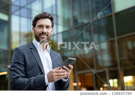 Professional man happily using a smartphone while standing outside a modern office building. Representing mobile communication. Business connectivity. And digital networking in a busy city environment Professional man happily using a smartphone while standing outside a modern office building. Representing mobile communication. Business connectivity. And digital networking in a busy city environment 138835784