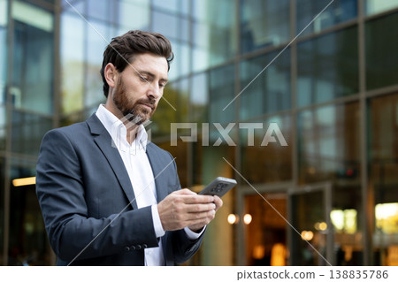 Businessman using smartphone for networking and corporate communication, standing outside a modern glass office building, representing mobile technology, connectivity, and urban professionalism 138835786