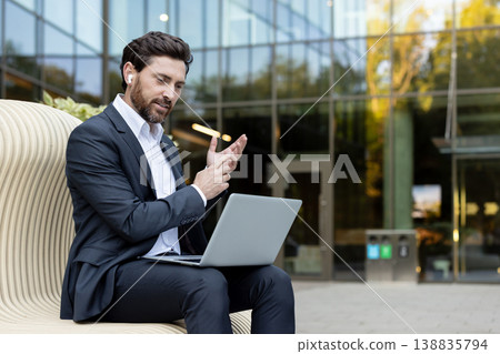 Businessman having a professional video call outdoors, using a laptop and wireless earbuds while sitting on a modern bench in front of a glass office building Businessman having a professional video call outdoors, using a laptop and wireless earbuds while sitting on a modern bench in front of a glass office building 138835794