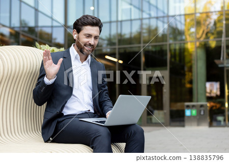 Smiling businessman wearing earbuds and suit, waving hand to laptop screen during an online video call for remote work or virtual meeting outside modern office building 138835796