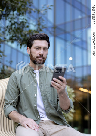Man with beard and casual clothes sitting on a stylish bench, intently reading or typing on his smartphone in an urban setting with a modern glass building in the blurred background Man with beard and casual clothes sitting on a stylish bench, intently reading or typing on his smartphone in an urban setting with a modern glass building in the blurred background 138835840