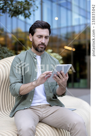 Man with beard and casual shirt sitting on a stylish bench, interacting with a digital tablet outdoors, focusing on work or communication in front of a modern building Man with beard and casual shirt sitting on a stylish bench, interacting with a digital tablet outdoors, focusing on work or communication in front of a modern building 138835842