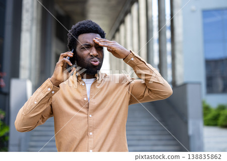 Young man feeling disappointed and frustrated, experiencing stress and headache while having a difficult phone conversation outside a modern city building, reacting to bad news Young man feeling disappointed and frustrated, experiencing stress and headache while having a difficult phone conversation outside a modern city building, reacting to bad news 138835862