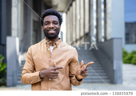 Smiling man standing outdoors in front of a modern building, happily pointing his fingers to the side, communicating options or showing the way forward Smiling man standing outdoors in front of a modern building, happily pointing his fingers to the side, communicating options or showing the way forward 138835870