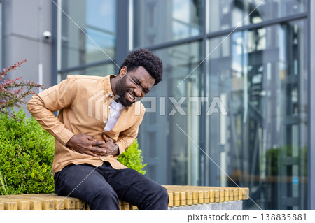 Young adult man experiencing severe abdominal cramps and discomfort while sitting on a bench in an urban environment, clutching his midsection with a pained expression 138835881