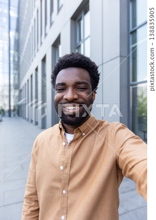 Confident young man with curly hair and beard smiling at camera outside a modern office building, casual entrepreneur portrait conveying success, friendliness, and urban optimism 138835905