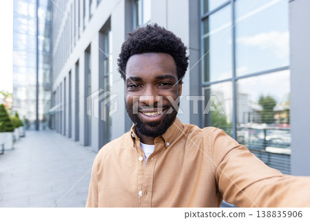 Young smiling black entrepreneur takes a confident outdoor selfie in a modern urban setting, candid headshot with office buildings in the bright daytime background, authentic and upbeat 138835906