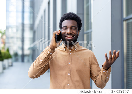 Smiling young professional walking through a modern city, talking and gesturing on his smartphone, confident and relaxed while handling business communication outdoors 138835907