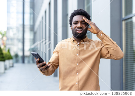 Young adult man experiencing headache and frustration while holding a phone outdoors, representing stress, financial problems, and business challenges Young adult man experiencing headache and frustration while holding a phone outdoors, representing stress, financial problems, and business challenges 138835908