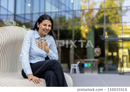 Indian woman clutching her chest while sitting on a modern bench outdoors, suffering from severe chest pain, a medical emergency indicating a heart attack or angina attack Indian woman clutching her chest while sitting on a modern bench outdoors, suffering from severe chest pain, a medical emergency indicating a heart attack or angina attack 138835918