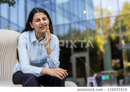 Young indian woman grimacing in pain, holding her cheek on a city bench, suffering from intense toothache and jaw discomfort while dressed in business casual outdoors Young indian woman grimacing in pain, holding her cheek on a city bench, suffering from intense toothache and jaw discomfort while dressed in business casual outdoors 138835919