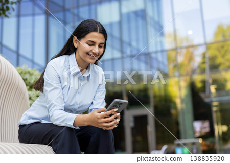 Young businesswoman smiling, sitting outdoors and using a mobile phone, representing remote work, digital communication, and modern technology in a corporate environment 138835920