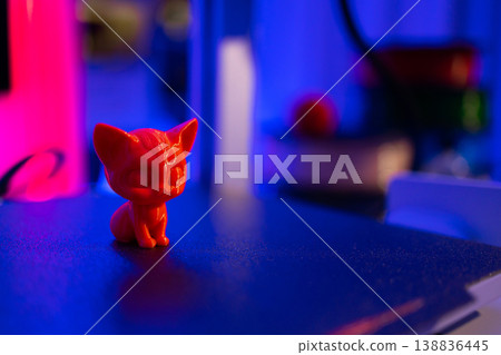 A small, bright red cat figurine sits on a dark surface illuminated by vibrant neon pink and blue lights, creating a futuristic atmosphere A small, bright red cat figurine sits on a dark surface illuminated by vibrant neon pink and blue lights, creating a futuristic atmosphere 138836445