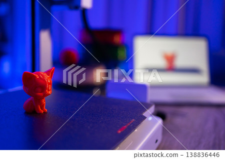 A small red 3D printed cat figurine sits on a 3D printer bed with a laptop displaying the printer software in the background A small red 3D printed cat figurine sits on a 3D printer bed with a laptop displaying the printer software in the background 138836446