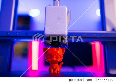 A 3D printer is actively creating a small, bright red cat figurine with intricate details under vibrant blue and pink lighting A 3D printer is actively creating a small, bright red cat figurine with intricate details under vibrant blue and pink lighting 138836449