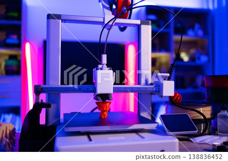 A modern 3D printer is actively creating an orange figurine on its build plate, illuminated by vibrant neon lights 138836452