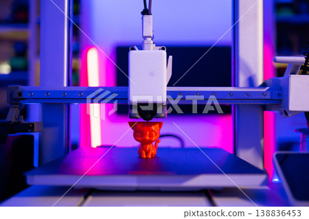 A 3D printer is actively creating a small, orange cat figurine with intricate details under vibrant blue and pink neon lighting 138836453