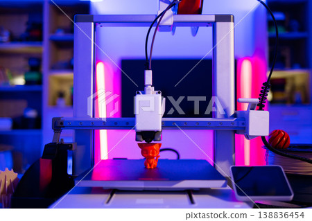 A modern 3D printer is actively creating a small orange cat figurine on its build plate, illuminated by vibrant neon lights A modern 3D printer is actively creating a small orange cat figurine on its build plate, illuminated by vibrant neon lights 138836454