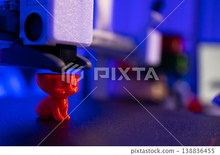 A 3D printer nozzle deposits molten plastic layer by layer to create a small, bright red cat figurine in a dimly lit workshop 138836455