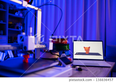 A 3D printer is actively creating a small orange cat figure, with spools of filament and a laptop displaying the 3D model nearby 138836457