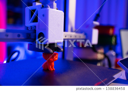 A close-up shot of a 3D printer in action, meticulously crafting a small, bright red cat figurine layer by layer under vibrant blue and pink lighting 138836458
